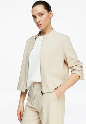Woman wearing beige tailored jacket and matching trousers, white shirt underneath, standing with one hand in pocket against white background.