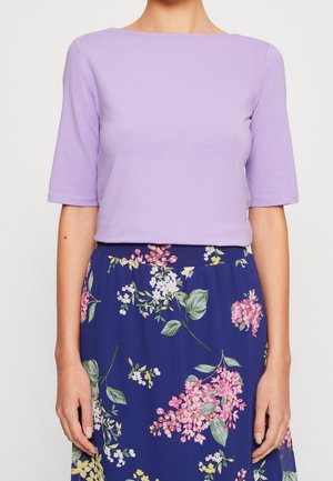 Kobieta ubrana w jasnofioletowy top z rękawami do łokcia, włożony do granatowej spódnicy z wysokim stanem ozdobionej różowo-białym kwiatowym wzorem.