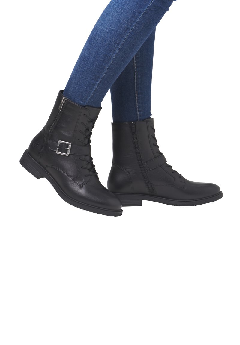 Schwarze Leder-Stiefeletten mit einer Schnürung an der Vorderseite, einem Reißverschluss an der Seite und einem Schnallen-Detail. Flache Laufsohle und strukturierte Oberfläche. Kombiniert mit blauen Jeans.