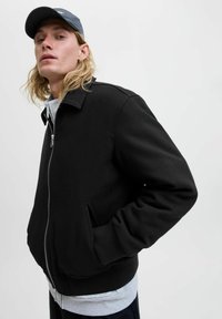 Zwarte zip-up jacket met een kraag, zijzakken en ribgebreide manchetten. Draagt over een grijze sweatshirt, weergegeven tegen een effen achtergrond.