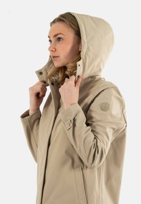 Chaqueta impermeable beige con capucha, que cuenta con botones a presión, bolsillos laterales y un logo en la manga. Textura suave y diseño aerodinámico.