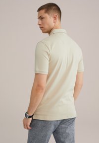 Camisa polo beige de mangas cortas, con cuello y canesú trasero. Combinada con pantalones azules texturizados y un reloj negro.