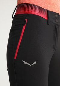 Pantalon de sport noir avec une ceinture rouge à motifs, une poche latérale zippée et un logo gris réfléchissant sur la cuisse. Texture de tissu lisse.