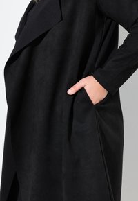Schwarzer Wildledermantel mit langen Ärmeln, oversized Kragen und Seitentasche. Hat eine glatte Textur und eine lockere Passform, mit einem leicht drapierten Design.