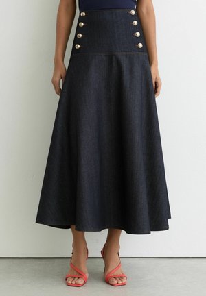 Kvinde iført en mørk denim midi-nederdel med høj talje og seks sølvknapper samt koralfarvede sandaletter med stropper og hæl, stående op ad en hvid væg.