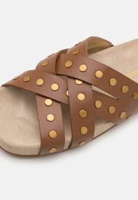 Καφέ λαστιχένια σανδάλια με διασταυρωμένες δερμάτινες λωρίδες, διακοσμημένα με χρυσά καρφιά, και κουμπωμένο μπεζ ποδοστάτη, σχεδιασμένα για άνεση και σταθερότητα.