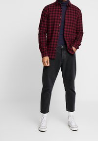 Rode en zwarte geruite flanellen shirt, gesloten tot aan de kraag, gecombineerd met donkere jeans en witte sneakers, gedragen over een marineblauwe coltrui.