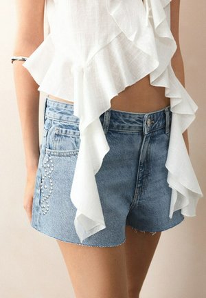 Top sans manches blanc à volants associé à un short en denim bleu clair avec des détails en strass sur la poche latérale.