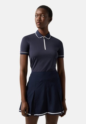 Femme portant un polo bleu marine à manches courtes avec des liserés blancs et une jupe plissée assortie avec une bordure blanche et des inserts en mesh.