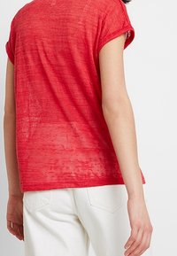 Top rosso a maniche corte con una leggera trasparenza e design con risvolto. Indossato con pantaloni bianchi, che mostrano una vestibilità rilassata e un tessuto liscio.