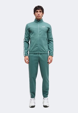Man standing wearing a green zip-up tracksuit with white stripes on sleeves and pants, white sneakers, neutral background.
