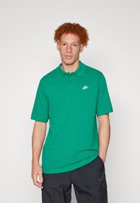 Vihreä puuvillapolo, jossa on klassinen kaulus ja lyhyet hihat. Vasemmassa rinnassa on valkoinen Nike-logo. Teksturoitu kangas, jossa on rento istuvuus.