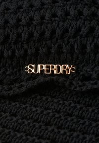 Gorro de punto negro con un patrón texturizado y un acento dorado del logo "SUPERDRY". Material de hilo suave con un ajuste ceñido.