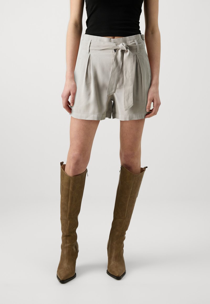Lichtgrijze shorts met een paperbag taille, voorzien van een strikriem, zijzakken en een relaxte pasvorm. Gecombineerd met bruine kniehoge laarzen.