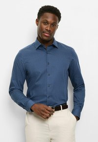 Marineblaues Hemd mit weißem geometrischem Muster, langen Ärmeln, Button-Down-Kragen und schwarzen Knöpfen; kombiniert mit hellfarbenen Hosen.