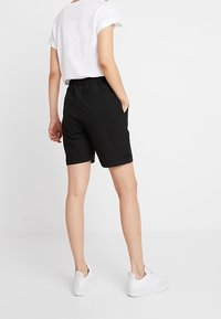 Persona che indossa pantaloni corti neri su misura, t-shirt bianca con maniche arrotolate e sneakers bianche, in piedi con le mani in tasca su uno sfondo bianco.