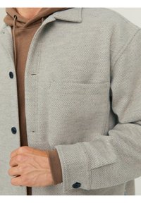 Jasnoszara koszula kurtka w jodełkę z kieszenią na piersi i czarnymi guzikami, noszona na brązowej bluzie z kapturem. Miękki, trwały materiał o teksturowanej powierzchni.