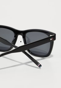 Ochelari de soare negri cu un design îndrăzneț și unghiular, lentile gri, finisaj lucios și un mic logo multicolor pe lateral.
