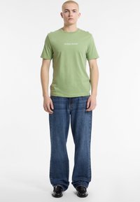 Zaļš kokvilnas T-krekls ar "GUESS JEANS" logotipu, kombinēts ar brīvā griezuma ziliem džinsiem un melnām kurpēm. Modelis stāv pret baltu fona.