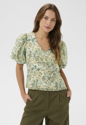 Nő világos, virágos mintás, rövid ujjú blúzban, dudorított ujjakkal és olívazöld nadrágban, kezét a zsebében tartva, egyszínű háttér előtt.