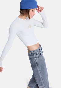 Λευκό ριμπ μακρυμάνικο τοπ με χρυσαφένιο έμβλημα, συνδυασμένο με άνετο γκρι τζιν. Το μοντέλο φοράει μπλε πλεκτό σκουφάκι.
