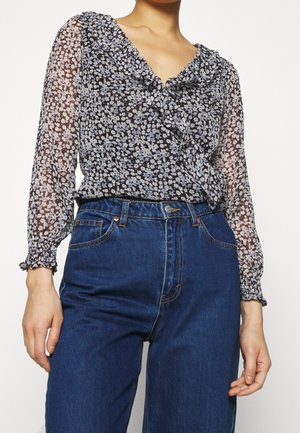 Vrouw draagt een doorschijnende bloes met lange mouwen en bloemenpatroon, voorzien van ruches, en gestopt in een hoog taille blauwe spijkerbroek.