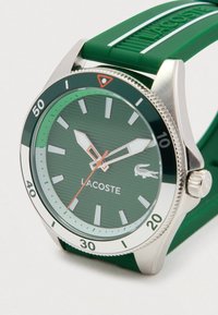 Roheline Lacoste käekell hõbedase äärisega, helendavate kätetega, punase sekundinäidikuga ja rohelise kummirihmaga, millel on Lacoste logo reljeef.