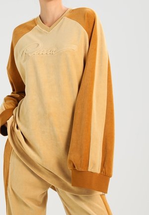 Fluwelen tweekleurige beige en bruine top met lange mouwen en ‘‘Puma‘‘-logo en bijpassende broek, gedragen door een staande persoon met hand in de zak.