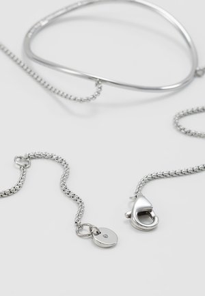 Collier chaîne en argent avec fermoir mousqueton et petite étiquette ronde comportant une pierre claire incrustée sur fond blanc.