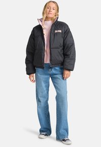 Jachetă puffer neagră cu căptușeală roz, având un logo pe piept, două buzunare frontale și manșete elasticate. Purtați cu blugi de culoare albastru deschis.