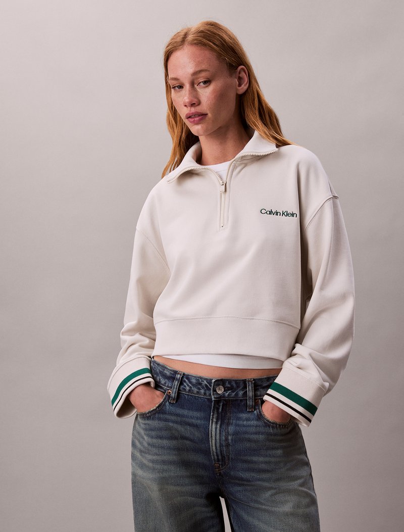 Bijela skratena dukserica s patentnim zatvaračem na ovratniku, koja sadrži zeleni "Calvin Klein" logotip. Ojačana zelenim i bijelim prugastim rukavima.
