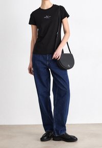 Person, der ein schwarzes Calvin Klein Jeans T-Shirt, lockere blaue Jeans, schwarze Lederloafers trägt und eine schwarze Calvin Klein Umhängetasche trägt.
