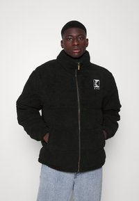 Jachetă neagră din fleece cu guler înalt, fermoar frontal și broderie cu logo. Prezintă o textură moale și manșete ribbate, asortată cu blugi din denim deschis.