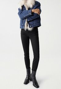 Person klädd i en blå denimjacka med vit päls, svarta skinnybyxor och svarta klackade ankelskos stående på vit bakgrund.