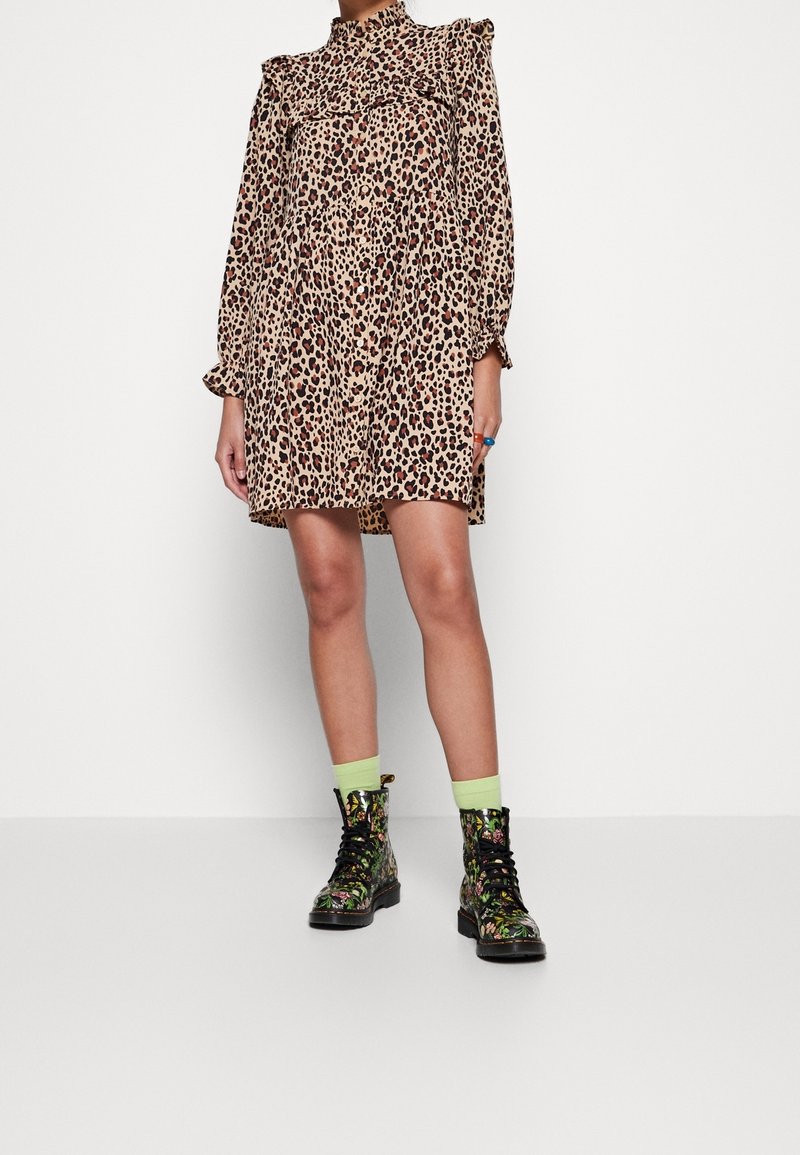 Personne portant une robe à manches longues imprimé léopard, des chaussettes vert néon et des bottes noires à lacets avec motif floral, debout sur un sol blanc uni.