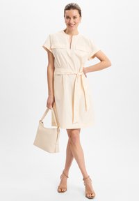 Frau lächelt, trägt ein knielanges cremefarbenes Kleid mit Taillengürtel, hält eine passende Handtasche und trägt beige Sandaletten mit hohen Absätzen.