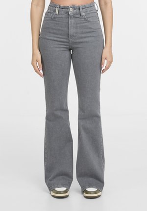 Kvinde iført grå, højttaljede, flared jeans med knap- og lynlås-lukning, kombineret med hvide og grønne sneakers, stående mod en hvid baggrund.