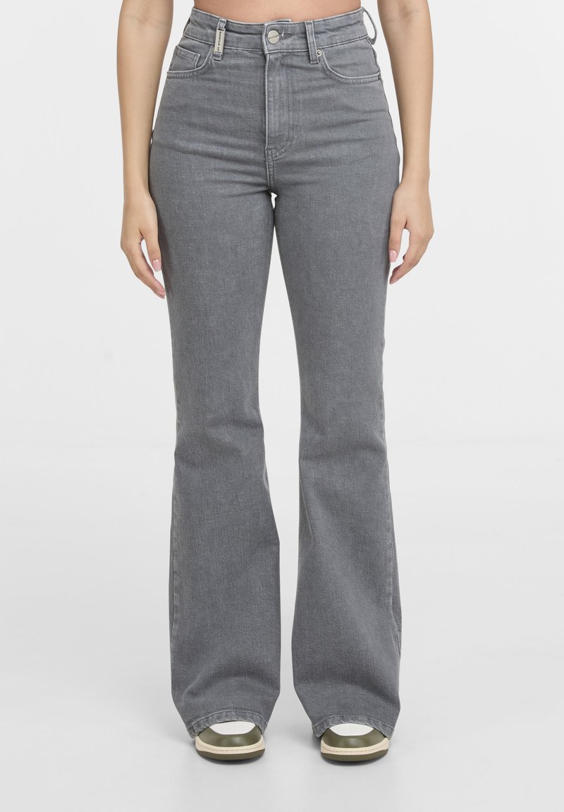 Femme portant un jean évasé gris taille haute avec une fermeture à bouton et zip, associé à des baskets blanches et vertes, debout devant un fond blanc.