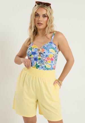 Blondynka ubrana w kwiecisty, niebiesko-żółto-różowy top bez rękawów z wycięciem na klatce piersiowej, żółte krótkie spodenki z wysokim stanem, okulary przeciwsłoneczne na głowie i złote bransoletki.