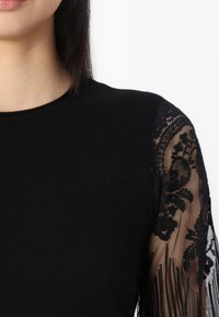 Svart topp med tettsittende liv, lange blondeermer med blomstermønster og en glatt tekstur. Flytende blonder gir kontrast.