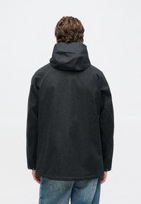 Man with wavy hair wearing a dark hooded jacket and light blue jeans, viewed from the back against a plain light background.