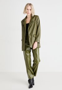 Olijfgroene neplederen blazer met een enkele knoop, gecombineerd met bijpassende textuurbroek. Zwarte enkelboots met hak maken de look af.