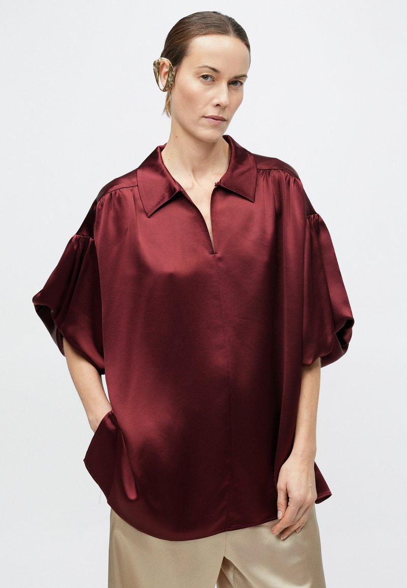 Woman wearing a burgundy satin blouse with wide sleeves and beige pants, standing against a plain white background.