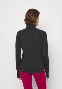 Schwarzes Langarmshirt mit hohem Kragen, ausgestattet mit strukturierten Details auf der Rückseite und einer schmalen Passform. Begleitet von bordeauxfarbenen Leggings mit seitlichen Streifen.