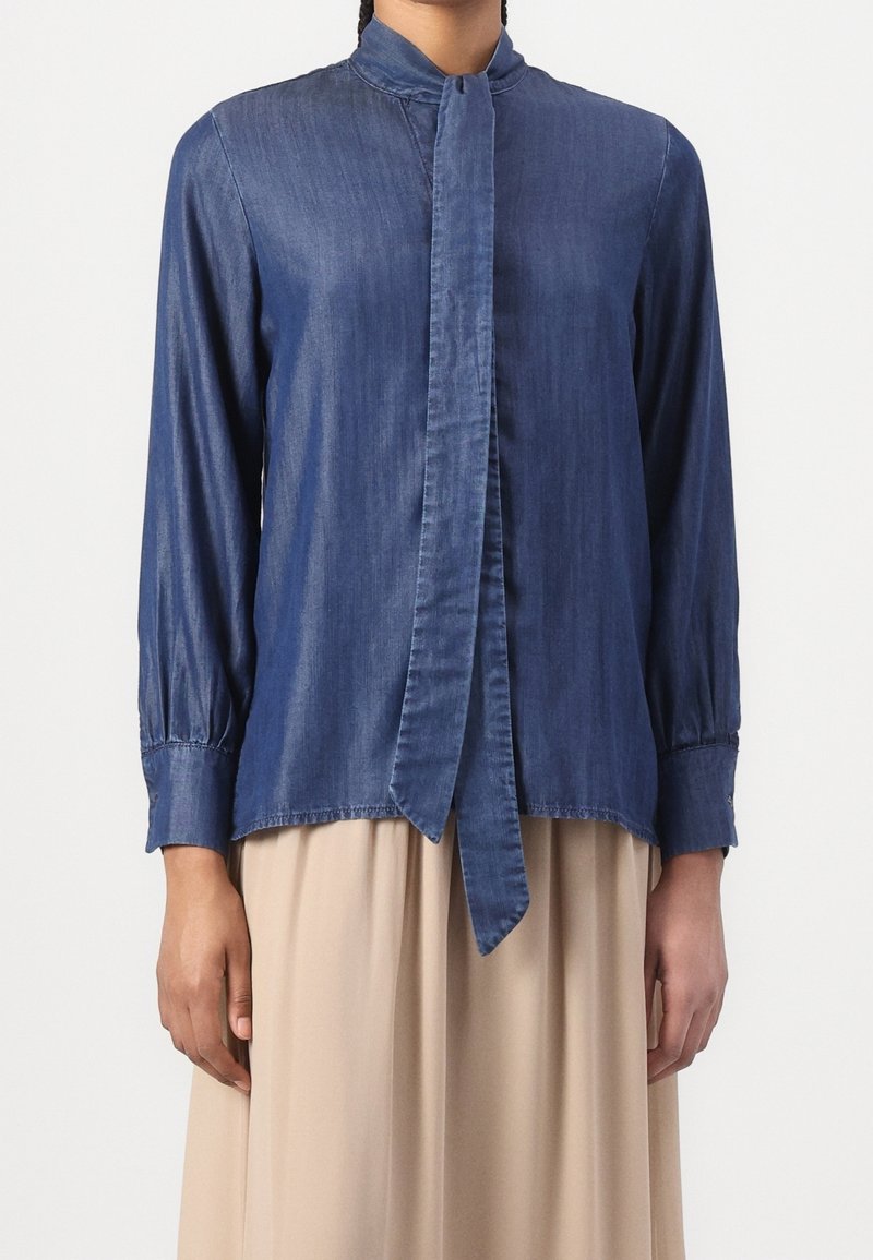 Femme portant une blouse bleue à manches longues avec un détail cravate, assortie à une jupe beige sur un fond uni.