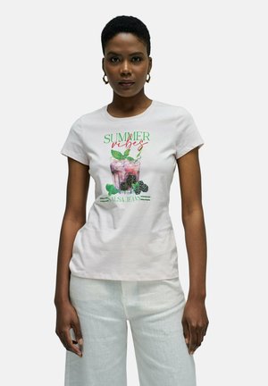 Mujer con camiseta blanca "Vibras de Verano" con un gráfico de bebida rosa y moras negras, combinada con pantalones blancos y pendientes de aro dorados.