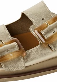 Guldmetallisk sandal med två stora transparenta spännen. Har slät yta, öppna sidor och en platt bas. Insidan är av beige tyg.