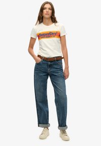 Jonge vrouw draagt een wit SuperDry T-shirt, een hooggetailleerde opgerolde blauwe spijkerbroek, een bruine riem en beige sneakers, staat met haar hand in haar zak.