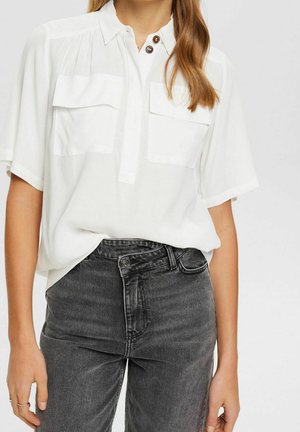 Kobieta nosząca luźną białą bluzkę z krótkim rękawem i dwoma kieszeniami na piersiach, włożoną do ciemnoszarych dżinsów z wysokim stanem i podwójnym zapięciem na guziki.