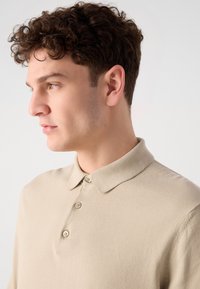 Polo beige con colletto, patta con tre bottoni e maniche corte; realizzato in un materiale morbido con una texture liscia.
